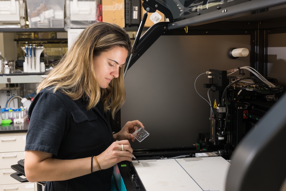 young woman uses tweezers to move small chips from a clear panel to a large machine as part of process to create a device of lung to brain chips.